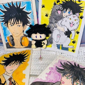 May include: A handmade black and yellow crochet doll stands in front of four watercolor illustrations of anime characters. The illustrations feature characters with black spiky hair and various facial expressions. The background includes cartoon images.
