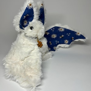 May include: A plush bat toy covered in white faux fur with large ears and wings lined in blue fabric featuring a pattern of white daisies and small bees. A small gold-toned charm shaped like a honeycomb hangs from the neck of the plush creature.