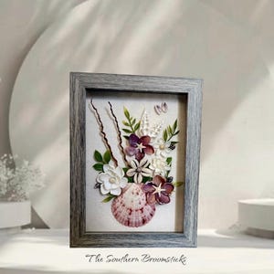 May include: A rectangular gray wood-grain frame containing a wall art piece featuring a bouquet of flowers crafted from various seashells. The arrangement includes white, purple, and brown shells, green leaves, and thin brown twigs on a light fabric.