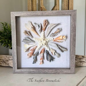 May include: A framed seashell art piece. A white starfish is surrounded by various seashells in a circular pattern on a white linen background. The frame is a light gray color, and the text "The Southern Broomsticks" is at the bottom.