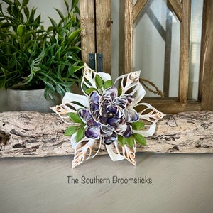 May include: A white seashell snowflake ornament with purple and white seashells in the center. The ornament is on a piece of driftwood and a white surface. The text "The Southern Broomsticks" is below the ornament.