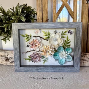 May include: A framed floral art piece with a light blue wooden frame. The artwork features a variety of handmade flowers in shades of white, peach, and teal, with green leaves and small butterfly accents. The text "The Southern Broomsticks" is at the bottom.