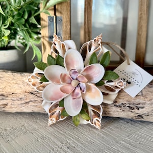 May include: A handcrafted seashell flower with a light pink and white color scheme. The flower is accented with green leaves and a small tag. The flower is mounted on a piece of driftwood.