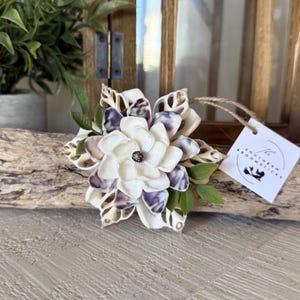May include: A handcrafted flower made from seashells, featuring white and purple petals with a dark center. Green leaves and a tag with the text "The Southern Broomsticks" are attached. The flower is displayed on a piece of driftwood.