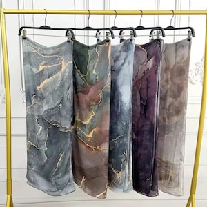 May include: Five silk scarves with a marbled design in shades of grey, green, purple, and brown, accented with gold veining. The scarves are hanging on a gold clothing rack.