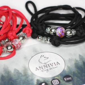Puede incluir: Dos collares o correas de paracord trenzado, uno rojo brillante y otro negro, cada uno con una cuenta de vidrio estampada central flanqueada por cuentas de metal plateado y nudos. Una tarjeta con el texto ANNIVIA Dog Wear es visible debajo.