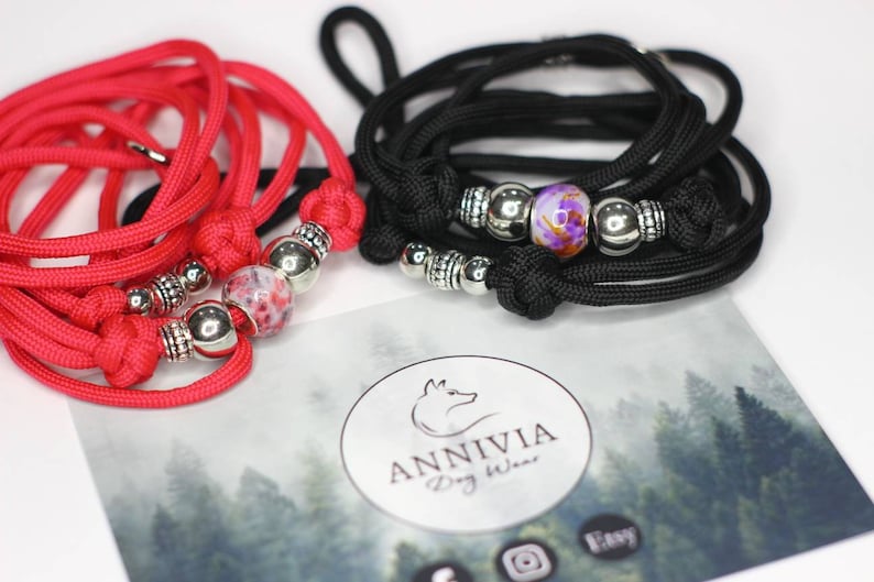 Puede incluir: Dos collares o correas de paracord trenzado, uno rojo brillante y otro negro, cada uno con una cuenta de vidrio estampada central flanqueada por cuentas de metal plateado y nudos. Una tarjeta con el texto ANNIVIA Dog Wear es visible debajo.