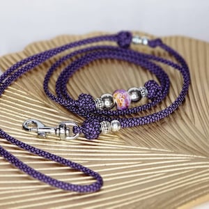 Puede incluir: Una correa de cuerda trenzada morada con un mosquet&oacute;n giratorio de metal plateado, cuentas decorativas plateadas y una cuenta central con un patr&oacute;n jaspeado en blanco y naranja. La correa est&aacute; hecha de cord&oacute;n resistente con nudos.