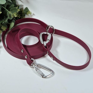 Puede incluir: Una correa para perro de color burdeos con dos clips plateados. La correa está enrollada y tiene un diseño plano y ancho.