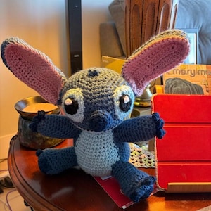 May include: A handmade crochet plush toy featuring a blue creature with large, upright ears that have pink inner sections. The figure has a light blue chest patch, dark blue limbs, and large black eyes with white highlights, crafted in a textured yarn.