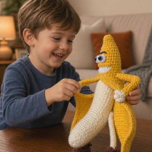 Peut inclure: Un personnage de banane crocheté avec un visage souriant, des yeux bleus et des chaussures marron. La banane est jaune avec un centre blanc et est tenue par un enfant. L'arrière-plan comprend un canapé et une lampe.