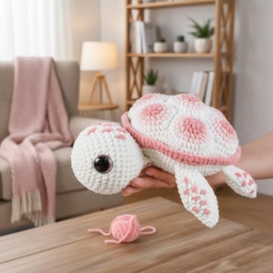 May include: A crocheted turtle plush toy featuring a white body with soft pink accents on the shell and flippers. The turtle has large, dark, glossy eyes and a textured yarn surface. A small ball of pink yarn sits on the wooden surface below.