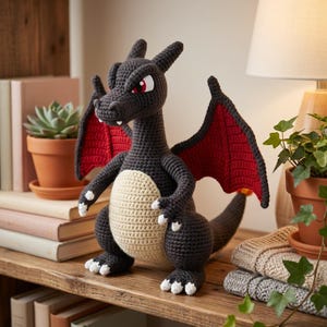 May include: A crocheted dragon figurine in shades of gray, red, and cream. The dragon has red wings, white accents on its feet and claws, and red eyes. It is displayed on a wooden shelf with books and plants.