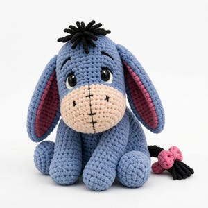 May include: A crocheted donkey plush toy featuring a periwinkle blue body, a light beige muzzle with black stitched details, and long ears with pink inner linings. The figure has a black yarn tuft on its head and a black tail with a pink bow.