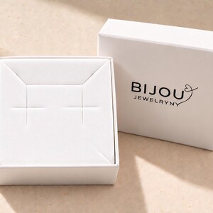 May include: A white square jewelry box with a removable lid featuring the black text BIJOU JEWELRYNY and a heart icon. The open base contains a white insert with two horizontal slits designed for holding earrings or small jewelry items.