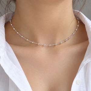 May include: A delicate silver chain necklace featuring small, evenly spaced round beads along the entire length. The necklace rests closely around the neck, showcasing a polished metallic finish that reflects light against the white shirt.