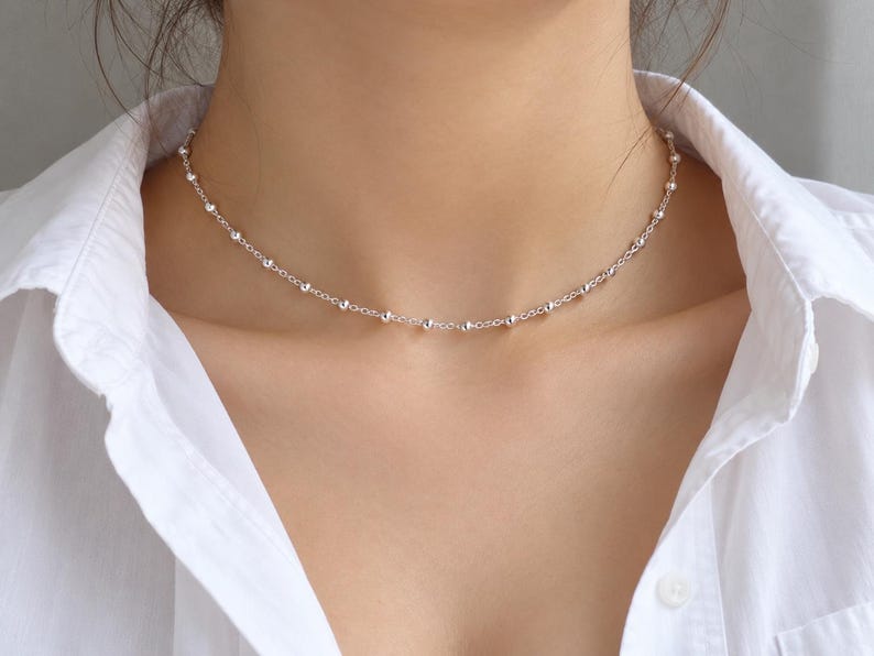 May include: A delicate silver chain necklace featuring small, evenly spaced round beads along the entire length. The necklace rests closely around the neck, showcasing a polished metallic finish that reflects light against the white shirt.