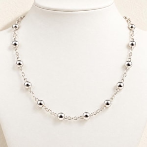 May include: A silver-toned necklace featuring a series of polished, spherical beads spaced evenly along a delicate metal chain. The necklace displays a consistent pattern of round, reflective beads connected by small, interlocking oval links.
