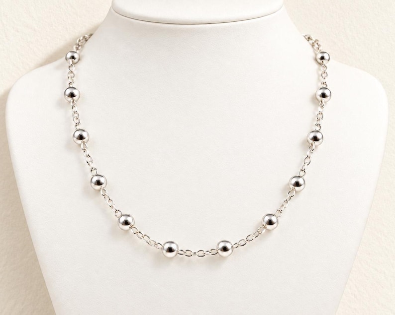 May include: A silver-toned necklace featuring a series of polished, spherical beads spaced evenly along a delicate metal chain. The necklace displays a consistent pattern of round, reflective beads connected by small, interlocking oval links.