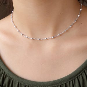 May include: A silver-toned chain necklace featuring evenly spaced small round beads along the length of the links. The necklace rests against the neck, showcasing a delicate metallic construction with a polished finish and a simple clasp.