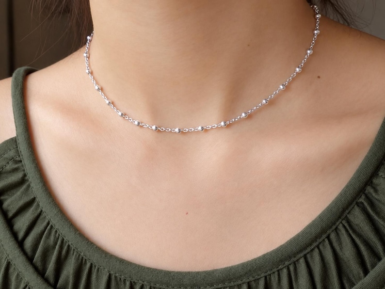 May include: A silver-toned chain necklace featuring evenly spaced small round beads along the length of the links. The necklace rests against the neck, showcasing a delicate metallic construction with a polished finish and a simple clasp.