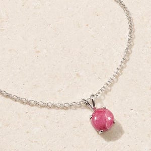 May include: A silver chain necklace featuring a round, polished pink gemstone pendant held in a four-prong setting. The pendant is centered on a delicate link chain, showcasing a smooth, vibrant pink stone with subtle white veining throughout.