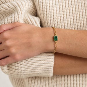 May include: A gold chain bracelet featuring a single rectangular green gemstone set in a gold frame. The bracelet consists of elongated oval links connected to the central stone setting, creating a delicate and refined piece of jewelry.