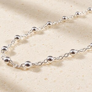 May include: A silver chain necklace featuring evenly spaced, polished spherical beads integrated into the links. The metallic chain has a bright, reflective finish and a delicate, uniform construction throughout its length.