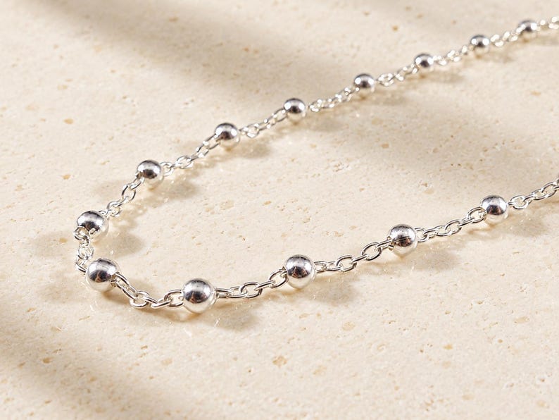 May include: A silver chain necklace featuring evenly spaced, polished spherical beads integrated into the links. The metallic chain has a bright, reflective finish and a delicate, uniform construction throughout its length.