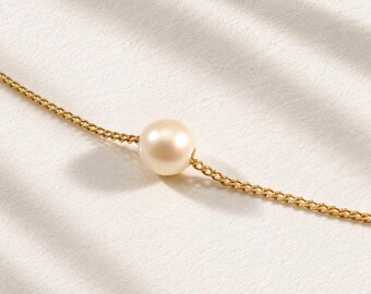 May include: A delicate gold chain necklace featuring a single, smooth, cream-colored pearl centered along the strand. The fine metal links of the chain connect to the pearl, creating a simple and elegant piece of jewelry with a polished finish.