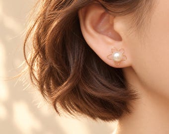 May include: A gold stud earring featuring a central white pearl surrounded by a thin, delicate gold wire frame shaped like a five-petaled flower. The earring is worn in the earlobe, showcasing the contrast between the pearl and gold.