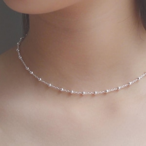 May include: A delicate silver chain necklace featuring evenly spaced small round beads along the entire length. The necklace rests closely around the neck, showcasing a polished metallic finish and a simple, refined link design.
