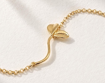 May include: A gold-toned bracelet featuring a delicate chain and a central charm shaped like a curved stem with two small, smooth leaves attached. The metal has a polished, reflective finish, and the design is connected to the chain links.