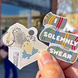 Puede incluir: Dos pegatinas de papel coloridas con temática de Harry Potter. La primera pegatina es una imagen pixelada de una casa con un esquema de colores amarillo, blanco y azul. La segunda pegatina es una bufanda a rayas gris y blanca con el texto "Juro solemnemente" y "que no estoy haciendo nada bueno".