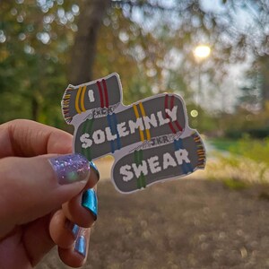May include: A gray and white sticker with the text "I solemnly swear" in a black font. The sticker is shaped like a folded scarf with red, yellow, and blue stripes.