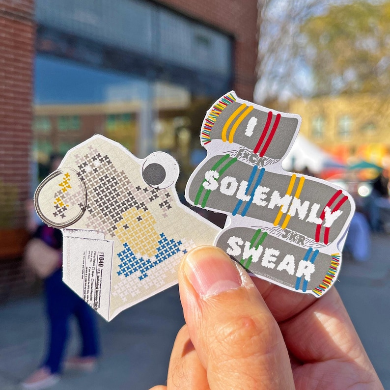May include: Two colorful paper stickers with a Harry Potter theme. The first sticker is a pixelated image of a house with a yellow, white, and blue color scheme. The second sticker is a gray and white striped scarf with the text "I solemnly swear" and "that I am up to no good".