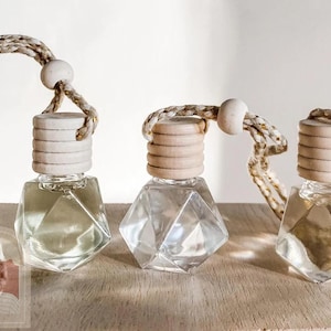 May include: Three clear glass bottles with a geometric design and wooden stoppers. Each bottle has a string attached to the stopper for hanging. The bottles are on a light brown wooden surface.