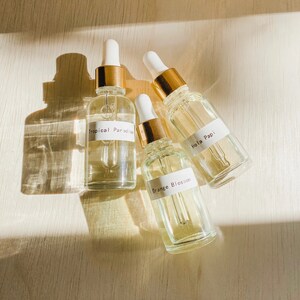 May include: Three glass bottles with gold caps and droppers. The bottles contain clear liquid and have white labels with the following text: "Tropical Paradise", "Orange Blossom", and "Hola Papi".