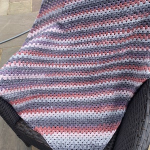 May include: A crocheted blanket with a striped pattern in shades of pink, gray, and white. The blanket is draped over a wicker chair.