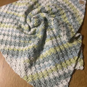 May include: A handmade crochet blanket in shades of white, light blue, and green. The blanket has a textured, patterned design and is folded over on a wooden surface. The blanket is soft and cozy.