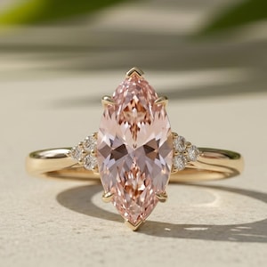 Könnte beinhalten: Ein goldener Ring mit einem großen pfirsichfarbenen Edelstein im Marquise-Schliff in der Mitte, flankiert von drei kleinen runden, klaren Steinen auf jeder Seite der Ringschiene. Der Metallring ist aus poliertem Gold und wirkt hell und glänzend.