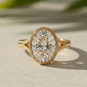 Op de afbeelding: Een gouden ring met een centrale ovaal geslepen heldere edelsteen in een kraalzetting. De ringband heeft een gespleten ontwerp dat taps toeloopt naar de centrale steen. De tekst Elista Jewel is op drie plaatsen rond de ring zichtbaar.