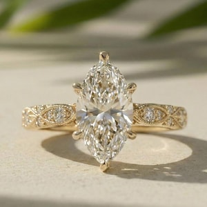 May include: A yellow gold ring featuring a large marquise-cut center diamond held by six prongs. The band is decorated with intricate metalwork and small round accent diamonds set along the sides, creating a detailed and textured appearance.
