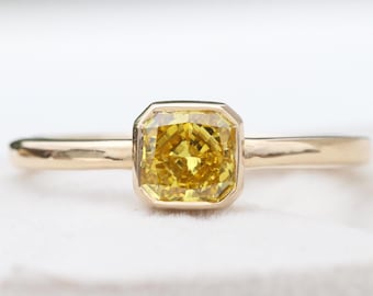 May include: A gold ring featuring a square-cut, vibrant yellow gemstone. The gemstone is set in a bezel setting, and the band is a simple, polished gold. The ring is displayed against a soft, white background, highlighting the ring's design.