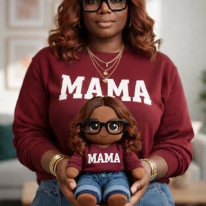 Op de afbeelding: Een vrouw draagt een bordeauxrode trui met het woord "MAMA" in witte letters en houdt een pop vast. De pop heeft een bruine huid, een bril en een bijpassende bordeauxrode trui met "MAMA" erop. De pop draagt een spijkerbroek.