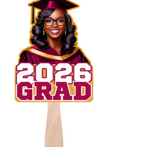 May include: A graduation fan featuring a smiling graduate in a maroon cap and gown. The fan displays the text "2026 GRAD" in maroon and white, with a gold border, and a wooden handle.