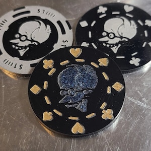 May include: Three poker chips featuring a cartoon character design. One chip is silver with black detailing and the text "$100". The other two chips are black with gold and white detailing. The chips are on a metallic surface.