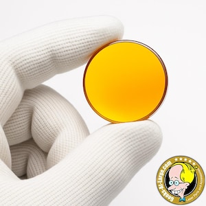 Puede incluir: Una lente redonda, transparente y teñida de naranja es sostenida por una mano enguantada. La lente tiene un borde marrón oscuro. La imagen incluye un logotipo con el texto "Geeks-At-Large.com" y "Laser Parts and Accessories."