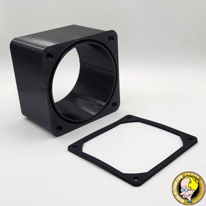 Puede incluir: Un componente negro de forma cuadrada con un interior cilíndrico y orificios de montaje. También hay una junta cuadrada negra separada con orificios coincidentes. Los artículos parecen estar impresos en 3D.