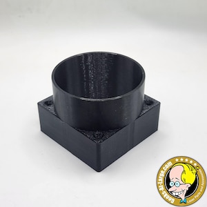 May include: A black, 3D-printed cylindrical object with a square base. The cylinder has a hollow interior and sits atop the square base, which has small holes in each corner. The object is likely a component for a larger device.